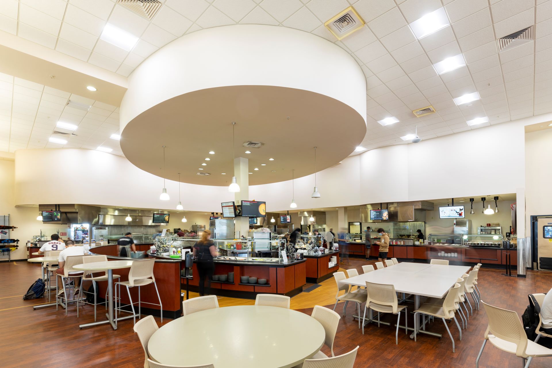 Wide interior view of a college dining hall with a large circular ceiling fixture, multiple food stations, and seating areas